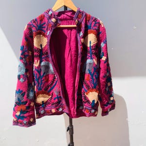 Op de afbeelding: Een magenta jasje met gedetailleerde bloemenborduurwerk in blauw, beige en rood. Het open jasje onthult een magenta voering. Het borduurwerk cre&euml;ert een textuur. Het jasje hangt aan een houten hanger. Afmetingen in centimeters.