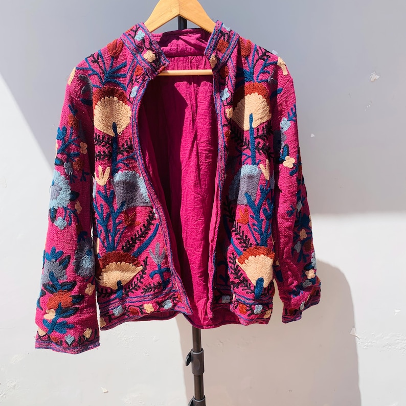 Op de afbeelding: Een magenta jasje met gedetailleerde bloemenborduurwerk in blauw, beige en rood. Het open jasje onthult een magenta voering. Het borduurwerk cre&euml;ert een textuur. Het jasje hangt aan een houten hanger. Afmetingen in centimeters.