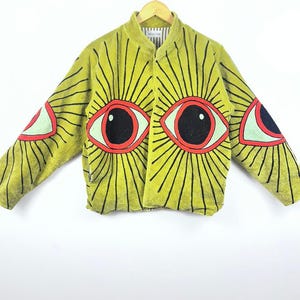 May include: A chartreuse jacket with a unique eye design. The jacket features large, stylized eyes with black radiating lines on the sleeves and front. The eyes have red outlines, black pupils, and light green irises. The jacket is hanging on a wooden hanger.