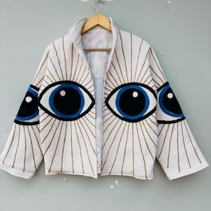 May include: White jacket with a unique eye design. The jacket features large blue and black eye appliqués on the front and sleeves, with gold-colored radiating lines. The jacket is open-fronted and has a quilted texture.