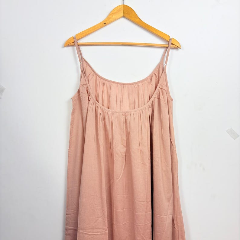 May include: A light pink spaghetti strap dress hanging on a wooden hanger. The dress has a scoop neckline and a gathered bodice. The fabric appears to be a soft, lightweight material, suitable for warm weather.
