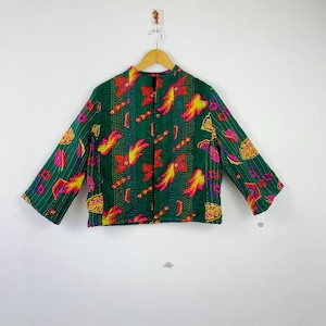 Puede incluir: Chaqueta verde con un colorido estampado floral, colgada de una percha de madera. La chaqueta presenta una parte delantera abotonada y mangas largas. El diseño floral incluye detalles rojos, amarillos y rosas.