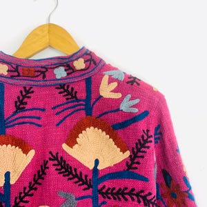 Op de afbeelding: Een close-up van een roze geborduurde jas met een bloemenmotief. De jas heeft cr&egrave;me, blauwe en rode bloemenborduursels. De jas hangt aan een houten hanger tegen een witte achtergrond.