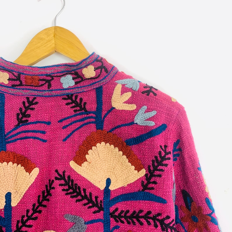 Op de afbeelding: Een close-up van een roze geborduurde jas met een bloemenmotief. De jas heeft cr&egrave;me, blauwe en rode bloemenborduursels. De jas hangt aan een houten hanger tegen een witte achtergrond.