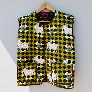 Puede incluir: Un chaleco acolchado con un patrón único. El chaleco presenta un diseño repetido de vacas blancas y flores de loto rosas sobre un patrón de diamantes verdes y negros. Tiene dos bolsillos delanteros y un ribete negro.