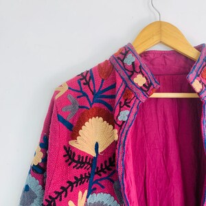 Op de afbeelding: Een felroze jas met ingewikkelde bloemenborduursels in blauw, rood en beige. De jas hangt aan een houten kleerhanger, wat het gedetailleerde ontwerp en de textuur benadrukt. De voering is effen magenta van kleur.
