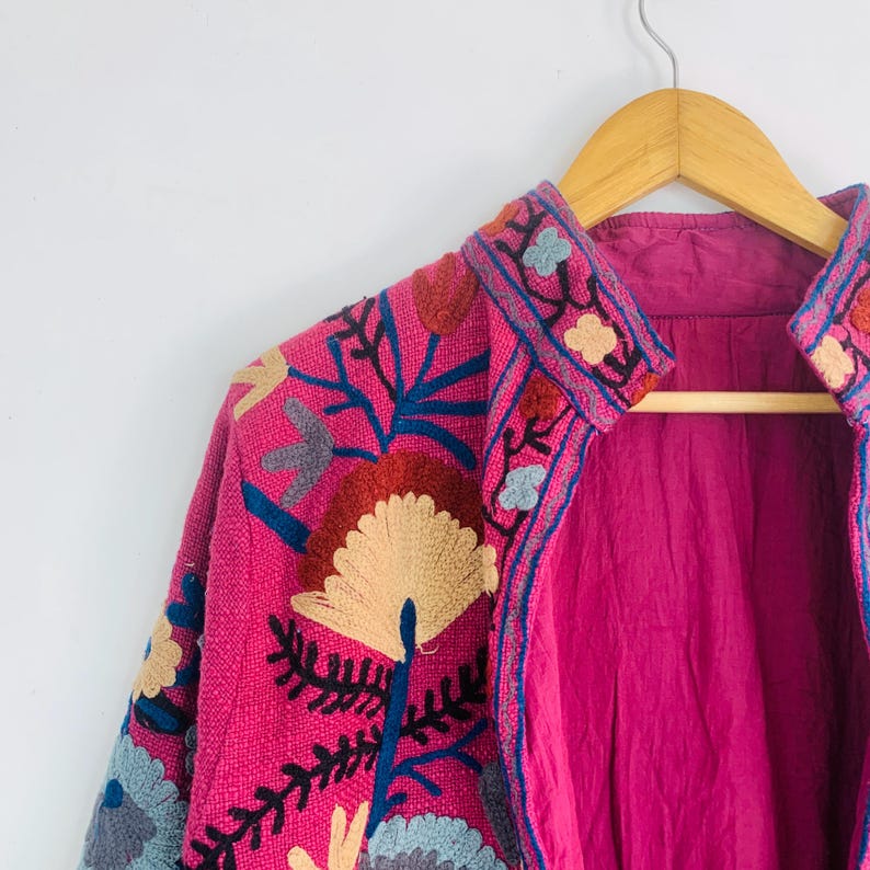 Op de afbeelding: Een felroze jas met ingewikkelde bloemenborduursels in blauw, rood en beige. De jas hangt aan een houten kleerhanger, wat het gedetailleerde ontwerp en de textuur benadrukt. De voering is effen magenta van kleur.