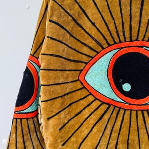 May include: Mustard-colored jacket with a large eye design. The eye features a black pupil, a light blue iris, and a red outline. Black lines radiate from the eye, creating a sunburst effect. The jacket's fabric appears to be velvet.