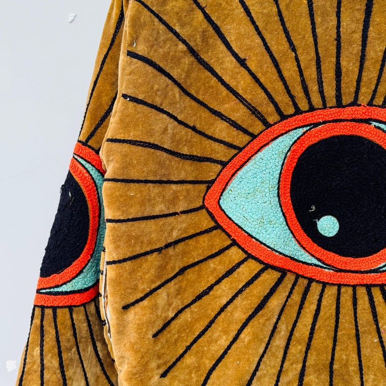 May include: Mustard-colored jacket with a large eye design. The eye features a black pupil, a light blue iris, and a red outline. Black lines radiate from the eye, creating a sunburst effect. The jacket's fabric appears to be velvet.