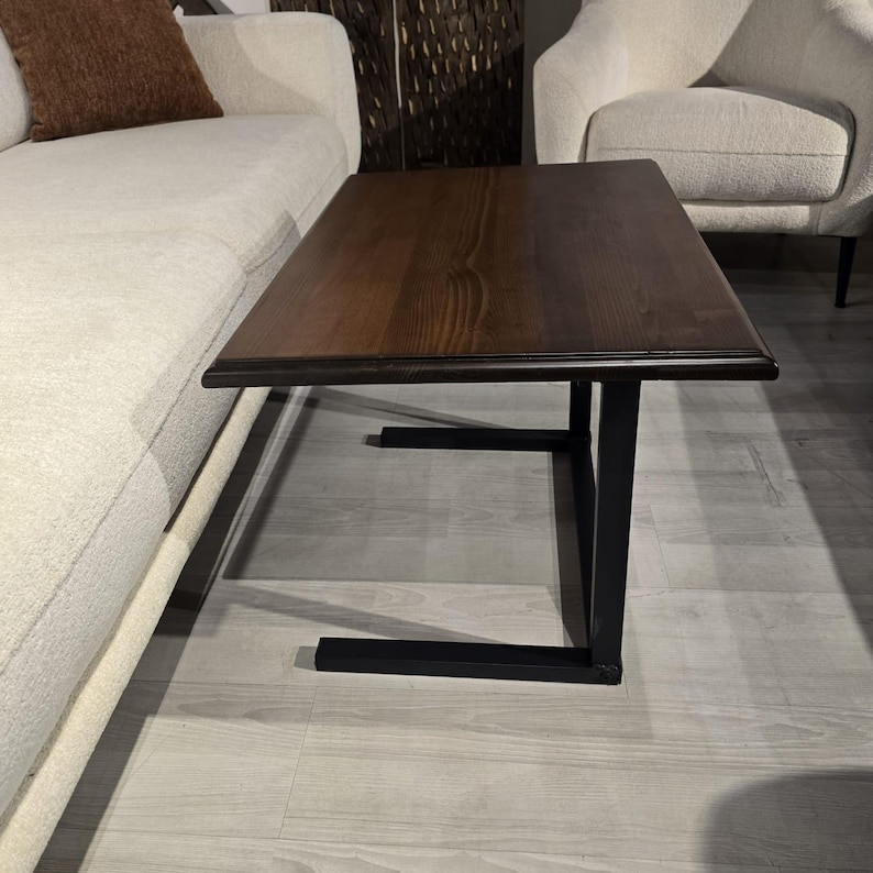 May include: A rectangular side table featuring a dark brown wooden top with a visible grain pattern and a black metal C-shaped frame base. The table is designed to slide over furniture, with a flat horizontal base and a vertical support leg.