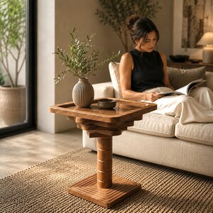 May include: A square wooden side table with a tiered, stacked design and a central cylindrical pedestal base. The table features a smooth, polished finish and sits on a square base with concentric grooved lines. A ribbed ceramic vase holds greenery.