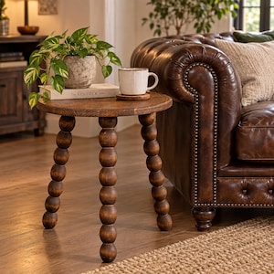 Puede incluir: Una mesa auxiliar redonda de madera con tres patas de diseño de esferas apiladas. La superficie de la mesa sostiene una taza de cerámica blanca sobre un posavasos de madera y una planta verde en maceta sobre el libro The Kinfolk Home.