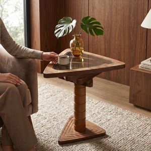 May include: A triangular side table crafted from dark wood with a central cylindrical pedestal base and a glass tabletop insert. The table features a three-sided base and a smooth, polished finish, providing a stable surface for a small ceramic cup.