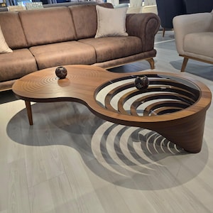 Op de afbeelding: Een unieke houten salontafel met een opvallend ontwerp. De tafel heeft een gebogen vorm met een glazen midden en een patroon van gesneden concentrische cirkels aan één kant. Twee decoratieve bollen liggen op de tafel. De tafel is ongeveer 122 cm lang.