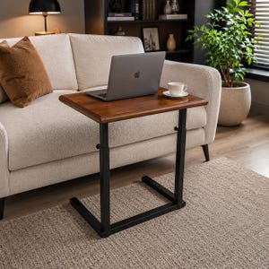 May include: A rectangular side table with a dark brown wooden top and a black metal C-shaped frame. The table features an adjustable height mechanism with visible knobs on the vertical supports, providing a stable base for a laptop and a cup.