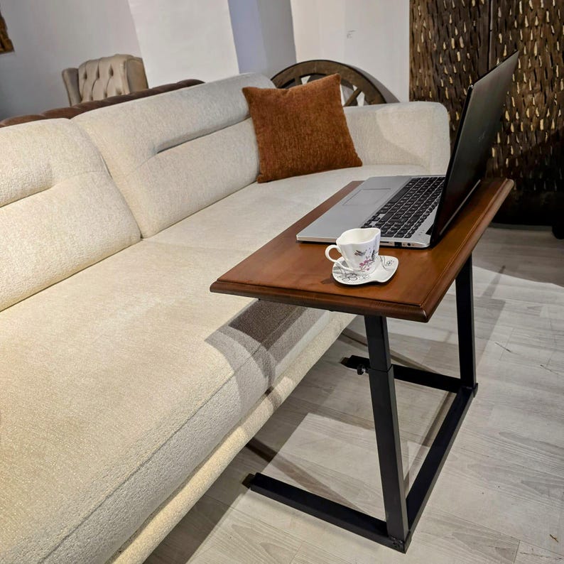 May include: A C-shaped side table with a dark wood top and a black metal frame. A silver laptop is open on the table surface next to a small white ceramic teacup and saucer with a delicate floral pattern. The table is positioned beside a sofa.