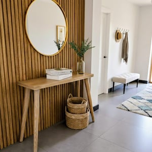 May include: A hallway scene featuring a wooden console table with a round gold mirror and a slatted wood wall. Two woven baskets sit below the table, and a bench and rug are visible in the background. A vase with greenery and books are on the table.