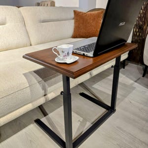 May include: A brown wooden laptop table with a black metal frame. A laptop and a teacup and saucer are on the table. The table is next to a beige sofa and a brown cushion.