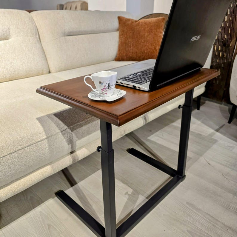 May include: A portable C-shaped side table with a rectangular wooden top and a black metal frame. A black Asus laptop sits on the surface next to a white ceramic teacup and saucer featuring a small Eiffel Tower graphic and floral accents.