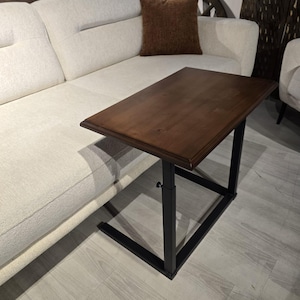 May include: A rectangular side table with a dark brown wooden top and a black metal frame. The table features an adjustable height mechanism with a small black knob on the side, designed to slide over the edge of a sofa or chair seat.