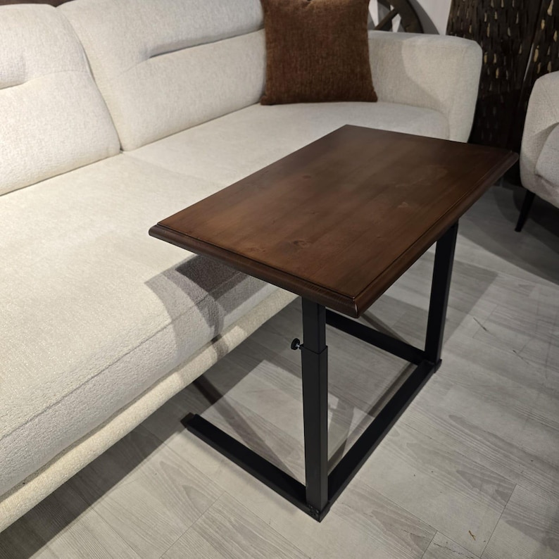 May include: A rectangular side table with a dark brown wooden top and a black metal frame. The table features an adjustable height mechanism with a small black knob on the side, designed to slide over the edge of a sofa or chair seat.