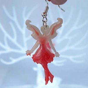Puede incluir: Pendiente con forma de hada con un gancho plateado. El hada tiene un cuerpo translúcido con detalles rojos en el vestido y las alas. Las alas están detalladas con vetas rojas. El pendiente está suspendido de un gancho plateado.