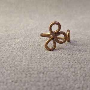 Puede incluir: Un anillo artesanal de color cobre con un diseño calado único. El anillo presenta una serie de formas en bucle, creando un patrón abstracto floral o en forma de nudo. El anillo está sobre un fondo neutro y texturizado.