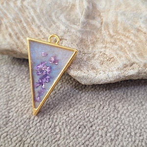 Op de afbeelding: Een goudkleurige, driehoekige hanger gevuld met lichtblauwe hars en paarse bloemaccenten. De hanger heeft een kleine lus aan de bovenkant voor een ketting. De achtergrond is een lichtgekleurde steen en een getextureerd oppervlak.
