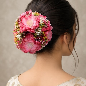 May include: A hair accessory featuring a cluster of artificial pink flowers, including large peony-like blooms, small white buds, and tiny green and yellow berries, arranged in a dense, rounded shape attached to dark hair in a bun style.