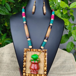 Pode incluir: Um conjunto de colar e brincos com um pendente quadrado de textura de bambu entrançado e um motivo central de um vaso de barro pintado e um fruto verde. O colar inclui contas cilíndricas de bambu e pequenas contas de madeira coloridas.
