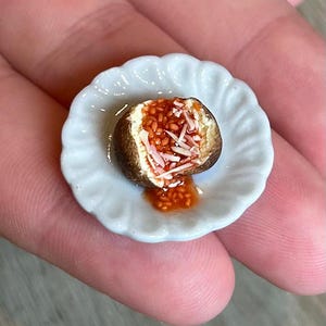 Könnte beinhalten: Miniatur-Gebäck, halbiert, mit brauner Kruste und roter Soßenfüllung, belegt mit weißen Streuseln, auf einem kleinen, weißen Teller mit gewelltem Rand. Eine kleine Menge Soße befindet sich auf dem Teller. Gehalten in einer Hand.