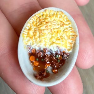 Puede incluir: Miniatura de pastel de pastor en un plato de cerámica blanco ovalado. El pastel tiene una corteza dorada y un relleno de carne, zanahorias y guisantes. El plato parece hecho a mano.
