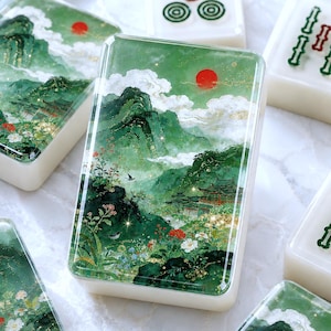 May include: A rectangular mahjong tile featuring a detailed green landscape illustration with mountains, a traditional building, white clouds, a red sun, and small birds. The design includes gold glitter accents and floral details.