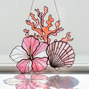 Puede incluir: Vidriera con una flor de hibisco, una concha y coral en tonos rosa, blanco y naranja. La obra de arte está suspendida por una cadena y está diseñada para colgarse en una ventana. El diseño tiene un tema costero.