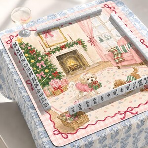May include: A rectangular mahjong mat featuring a festive Christmas living room illustration with a fireplace, a decorated tree, and two dogs. The mat is bordered by white mahjong tiles and includes two clear glass cups on the corners of the table.