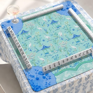 Op de afbeelding: Een vierkante mahjongmat met een lichtblauwe achtergrond voorzien van een patroon van witte kraanvogels, bamboe en blauwe pagodes. De mat heeft blauwe paraplumotieven in de hoeken en is omringd door witte mahjongstenen met zwarte tekens.