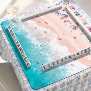 Könnte beinhalten: Eine quadratische Mahjong-Matte mit einer Draufsicht auf einen Strand mit türkisfarbenen Meereswellen und rosafarbenem Sand. Die Matte ist von weißen Mahjong-Steinen in Reihen umgeben, wobei einige Steine schwarze chinesische Schriftzeichen zeigen.