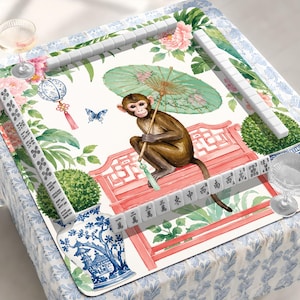 May include: A mahjong mat featuring a brown monkey holding a green parasol while sitting on a pink bench. The design includes pink peonies, a blue and white lantern, a butterfly, and a blue and white vase. White mahjong tiles are arranged around the edges.