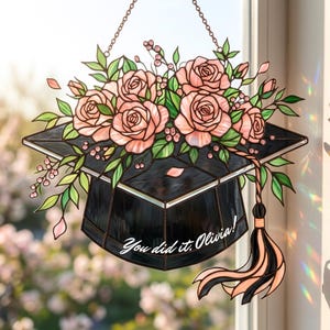 May include: Stained glass graduation cap with a bouquet of pink roses and green leaves. The cap is black with a tassel and chain for hanging. Text reads "You did it, Olivia!"