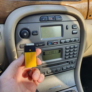 May include: A rectangular electronic device with a yellow lower section and a black upper section, featuring a small circular light and a letter J embossed on the front, held in front of a car dashboard with a digital clock displaying 14:01.