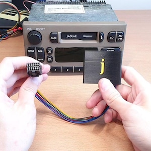 Könnte beinhalten: Ein Autoradio mit Jaguar-Logo, ein schwarzer Stecker und eine schwarze Blende mit gelbem "j" werden vor dem Radio gehalten. Der Stecker hat Drähte in verschiedenen Farben.