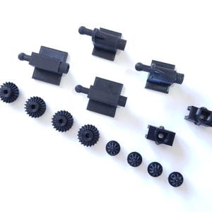 May include: A collection of black 3D printed mechanical components including three rectangular mounts with ball-joint ends, four small circular gears with teeth, two square-shaped housing brackets, and four tiny circular gear-like pieces.