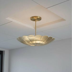 Op de afbeelding: Een messingkleurige, bloemblad-vormige hanglamp die aan het plafond hangt. De lamp heeft een centrale steel en een cirkelvormige plafondbevestiging. De lamp bevindt zich in een kamer met witte muren en een wit plafond.
