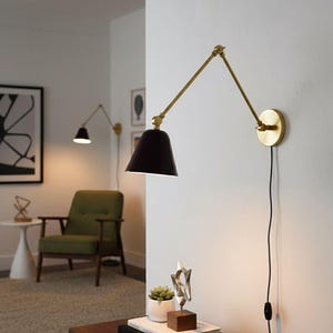 Op de afbeelding: Een zwarte en messingkleurige wandlamp met een verstelbare arm. De lamp heeft een conische kap en een ronde basis. De lamp is aan een witte muur bevestigd. Een tweede lamp is op de achtergrond zichtbaar. De lamp is aangesloten op een zwart netsnoer.