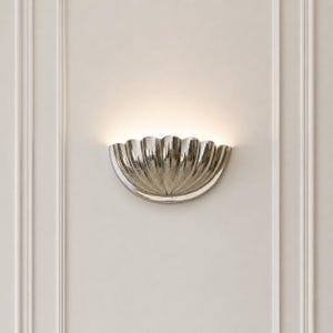 May include: A semi-circular wall sconce with a metallic silver finish and a scalloped, shell-like design. The fixture emits a soft, warm glow from its upper edge, casting light upwards against the wall surface to create a subtle, ambient illumination.