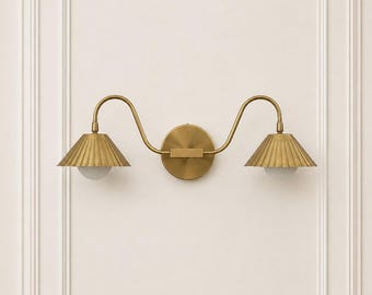 May include: A double-arm wall sconce finished in brushed brass. The fixture features a central circular mounting plate with two curved arms extending outward, each supporting a pleated, cone-shaped metal shade and a small white glass bulb.