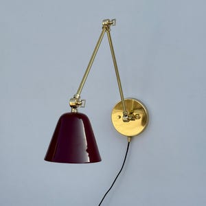 Op de afbeelding: Een wandlamp met een bordeauxrode kap en een verstelbare arm in messingkleur. De lamp heeft een ronde basis en een zwart snoer. Het ontwerp is modern en minimalistisch, geschikt voor diverse interieurstijlen.