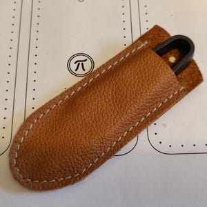 Puede incluir: Una funda de cuero marrón con costuras blancas contiene un objeto de metal negro. La funda tiene una superficie texturizada y está sobre una superficie blanca con un símbolo Pi.