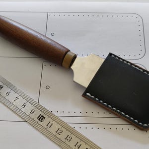 May include: A leathercraft knife with a dark wooden handle and a stainless steel blade, partially sheathed in a black leather case with white stitching. A metal ruler is in the foreground.