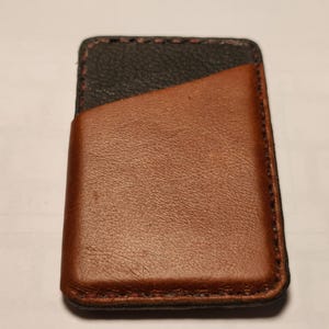 Könnte beinhalten: Handgefertigter Leder-Kartenhalter in zweifarbigem Design. Der obere Bereich ist schwarz, während die Vordertasche und der Körper warmbraun sind. Die Kanten sind mit einem Kontrastfaden genäht, was ein langlebiges Accessoire ergibt.